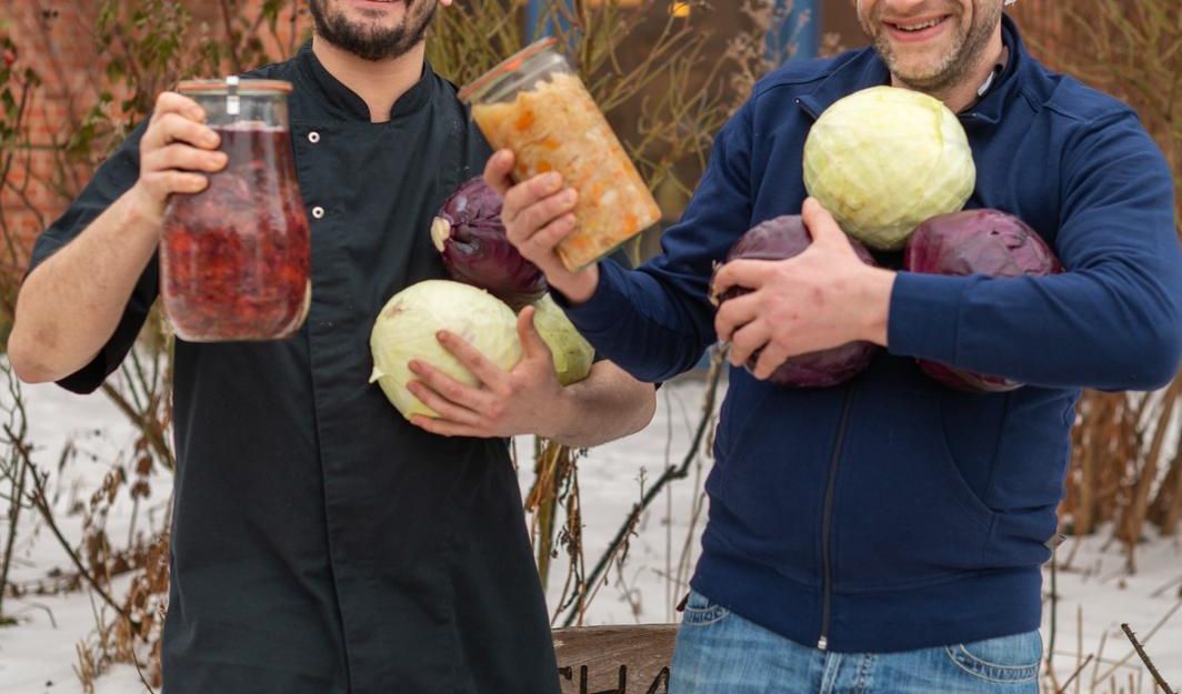 Zwei Männer halten Gläser mit fermentiertem Gemüse und Kohlköpfe in den Armen, vor einem Backsteingebäude im Schnee.