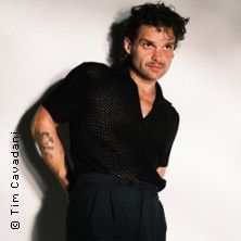 Ein Mann in schwarzem Hemd und dunkler Hose steht vor einem hellen Hintergrund.