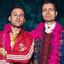 Zwei Männer in auffälliger Kleidung, einer in einem roten Trainingsanzug, der andere in einem historischen Kostüm mit pinkem Federboa.