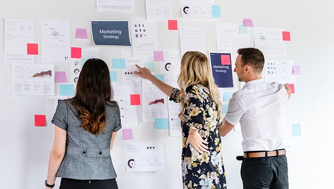 Es sind drei Personen zu sehen, die vor einer Wand mit Post-Its stehen