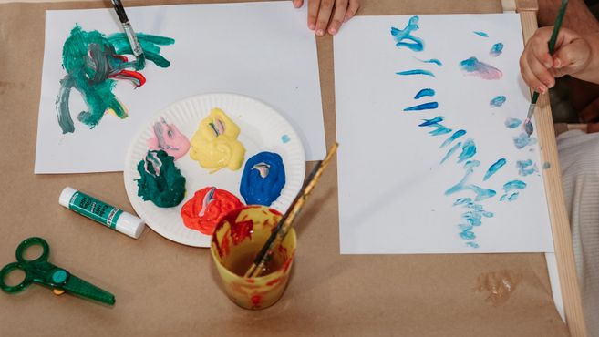 Kinder malen mit Pinseln auf Papier; auf einem Teller liegen bunte Farbtupfer, daneben Schere und Farbtube.