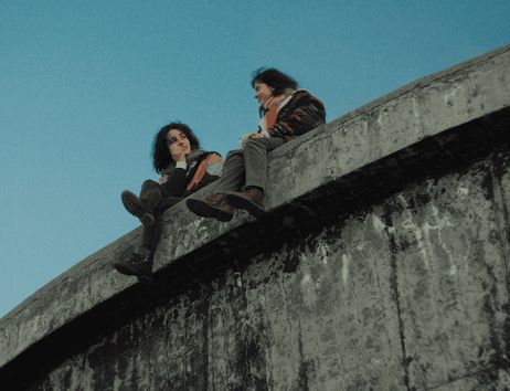 Zwei Personen sitzen auf dem Rand einer großen Betonstruktur vor blauem Himmel, passend zum Titel „37. Hamburg International Queer Film Festival - Miss Carbón (Queen of Coal) | Eröffnungsfilm“.