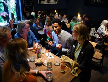 Menschen sitzen in kleinen Gruppen an Tischen und tauschen sich in einem Café über verschiedene Themen aus. Im Vordergrund ist eine Gesprächsrunde zu sehen.