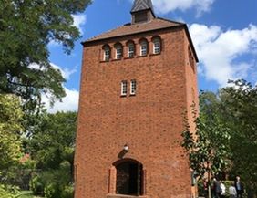 Backsteinturm mit Spitzdach vor blauem Himmel und Bäumen.