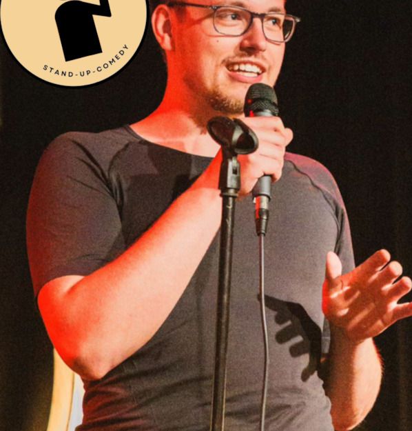 Ein Mann mit Brille hält ein Mikrofon auf einer Bühne. Oben links ist ein Logo mit der Aufschrift 'Uelzen-Mic Stand-up-Comedy'.