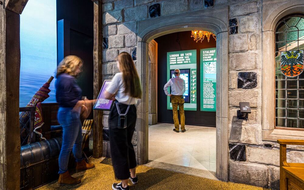 Foto in die Ausstellung. Im vorderen Raum schauen sich zwei Frauen eine Ausstellungstafel an. Durch einen Torbogen aus Stein schaut man in den nächsten Ausstellungsraum. Hier steht ein Mann vor einer Ausstellungstafel.