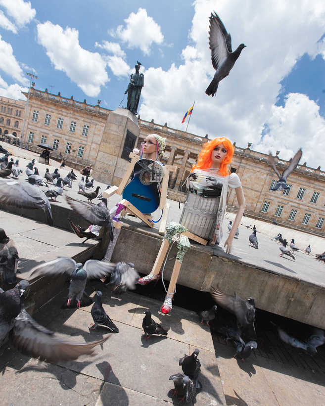 The image shows two mannequin-like, human figures assembled from mixed materials and mechanical parts, seated on a stone ledge in a public square. Pigeons surround them and fly through the frame. Historic architecture, a statue, and a bright blue sky form the background.