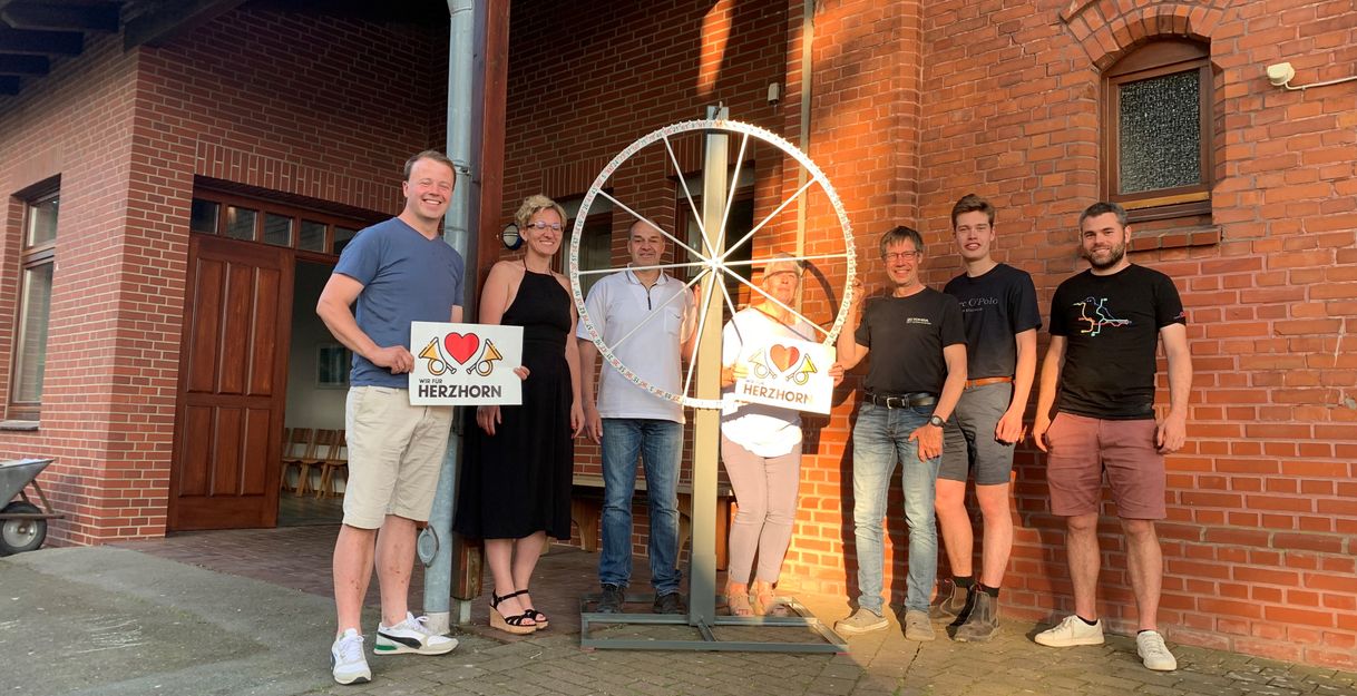 Eine Gruppe von sechs Personen steht vor einem Backsteingebäude neben einem großen Rad mit einem Schild, auf dem 'Herzhorn' steht.
