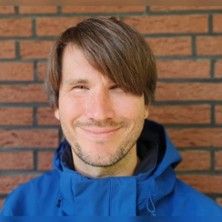 Ein Mann mit braunen Haaren und blauer Jacke lächelt vor einer Ziegelwand.