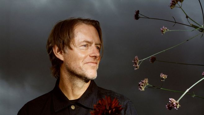 Ein Mann mit mittellangem Haar und Bart schaut lächelnd zur Seite. Im Vordergrund sind Blumen zu sehen, der Hintergrund ist dunkel und wolkig.