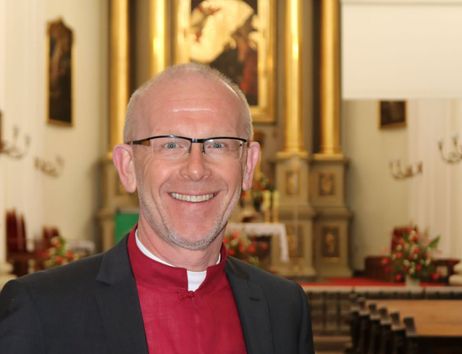 Ein Mann in einem roten Hemd und schwarzem Anzug steht lächelnd in einer Kirche mit einem goldenen Altar im Hintergrund.