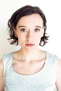 Porträt einer Frau mit kurzen braunen Haaren und grauem Oberteil vor weißem Hintergrund.