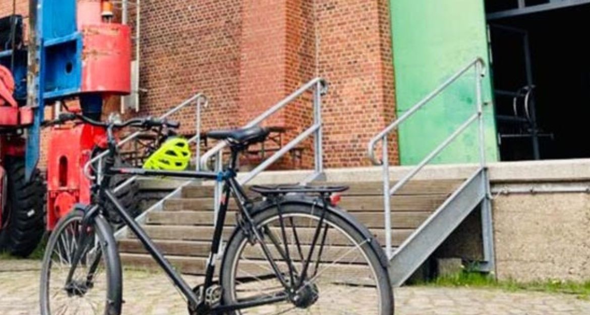 Ein Fahrrad steht vor einem Backsteingebäude mit einer grünen Tür und einer Treppe. Ein gelber Helm liegt auf dem Fahrradlenker.