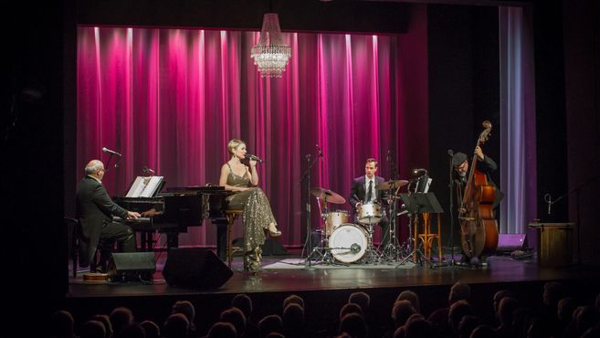 Eine Sängerin und ihre Band treten auf einer Bühne mit pinkem Vorhang auf. Ein Pianist, ein Schlagzeuger und ein Kontrabassist begleiten sie.