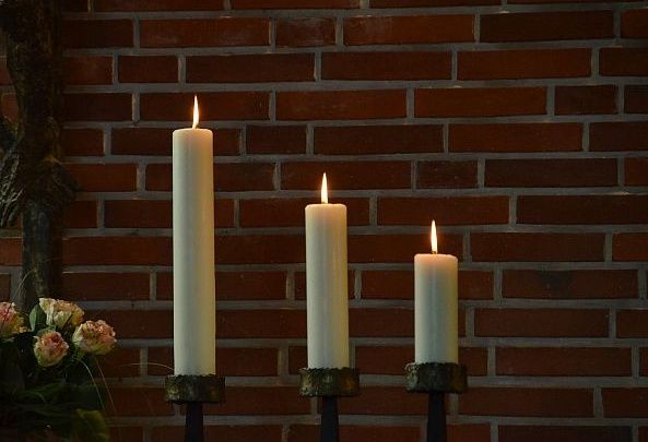 Drei brennende Kerzen vor einer Ziegelwand in einer Kirche.