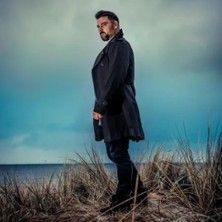 Person in dunklem Mantel steht am Strand mit bewölktem Himmel im Hintergrund.