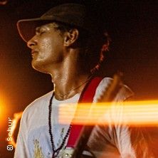 Musiker mit Gitarre und Mütze auf der Bühne, beleuchtet von warmem Licht.