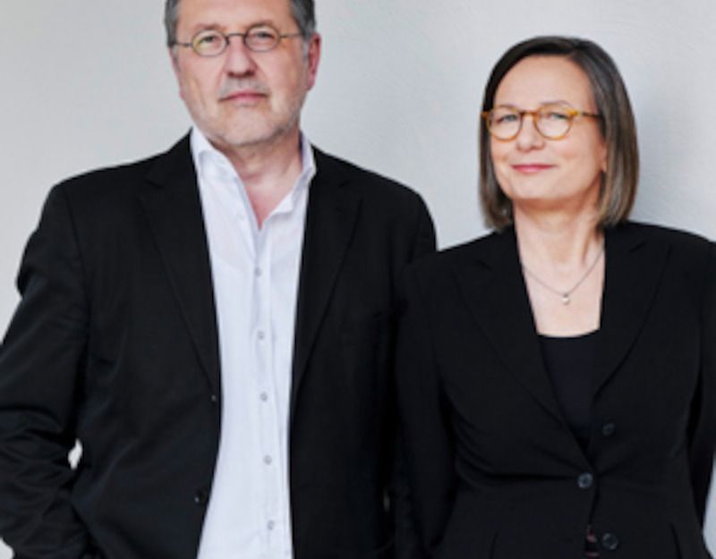 Ein Mann und eine Frau in formeller Kleidung stehen nebeneinander vor einem neutralen Hintergrund.