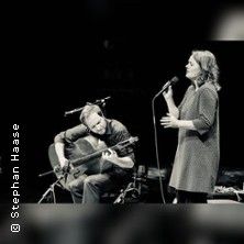 Ein Musiker spielt Gitarre, während eine Sängerin daneben steht und singt. Beide sind in einem Konzertsetting auf einer Bühne.