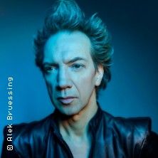 Porträt eines Mannes mit hochgestyltem Haar und Lederjacke vor blauem Hintergrund.