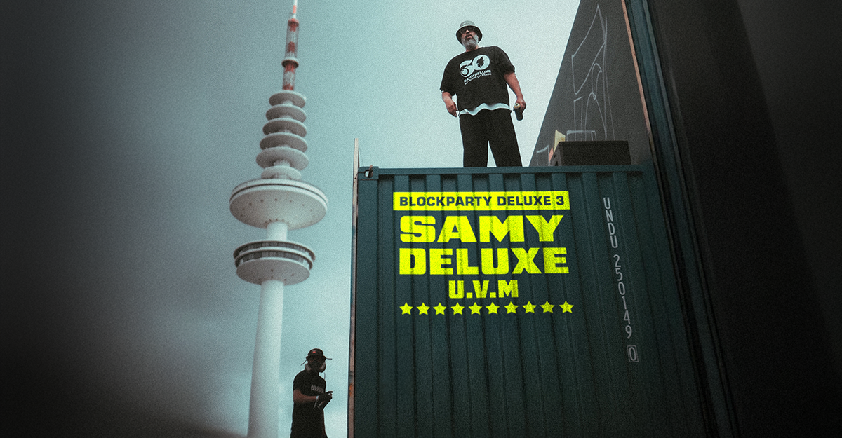Ein Mann steht auf einem Container mit der Aufschrift 'Blockparty Deluxe 3 Samy Deluxe U.V.M'. Im Hintergrund ist ein hoher Fernsehturm zu sehen.
