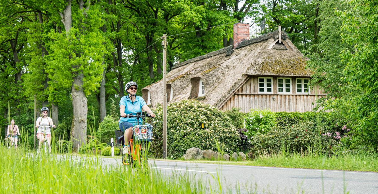 Drei Radelnde in grüner Natur, im Hintergrund ein reetgedecktes Haus