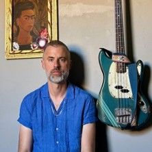 Ein Mann in einem blauen Hemd sitzt neben einer hängenden E-Gitarre. Im Hintergrund ist ein Gemälde von Frida Kahlo zu sehen.