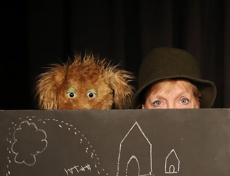 Eine Person mit Hut und eine zottelige Hunde-Handpuppe schauen hinter einer Tafel mit Kreidezeichnungen eines Hauses und eines Baumes hervor.