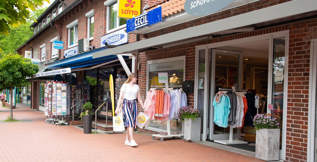 Eine Frau geht an Geschäften vorbei, darunter ein Bekleidungsgeschäft und ein Lottoladen, auf einer Einkaufsstraße.
