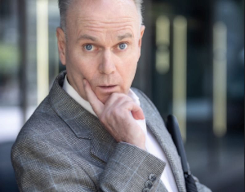 Ein Mann in einem grauen Anzug schaut überrascht in die Kamera und stützt sein Kinn mit einem Finger.