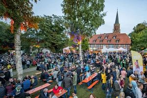 Menschenmenge bei einem Bierfest im Freien mit Bänken und einem historischen Gebäude im Hintergrund.