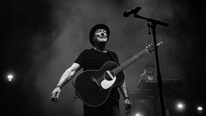 Ein Musiker mit Gitarre und Hut auf einer Bühne, im Hintergrund ein Keyboardspieler.