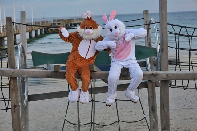 Zwei Personen in Osterhasenkostümen sitzen auf einem Klettergerüst am Strand mit einem Steg im Hintergrund.