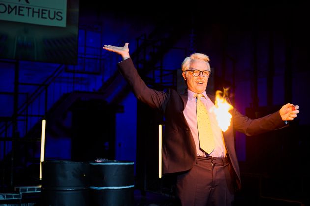 Schauspieler Samuel Weis im Anzug, einer grauen Perücke und Krawatte entzündet begeistert und effektvoll ein Stück Papier.