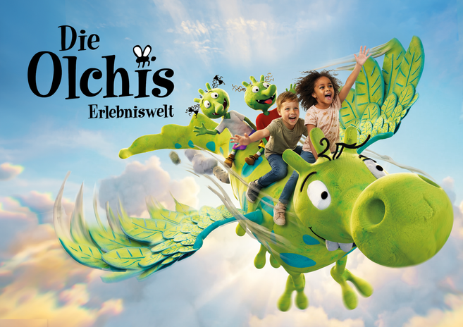 Zwei Kinder fliegen gemeinsam mit zwei grünen Olchi-Figuren auf dem großen grünen Drachen Feuerstuhl durch einen blauen Himmel voller Wolken.