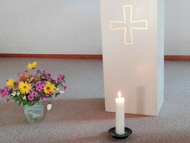 Ein Blumenstrauß und eine brennende Kerze vor einem weißen Podest mit Kreuzsymbol.