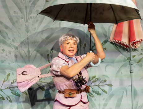 Frau mit kurzem grauem Haar hält einen geöffneten Regenschirm und trägt ein rosa Kleid mit passender Tasche vor einer Wand mit Pflanzenmuster