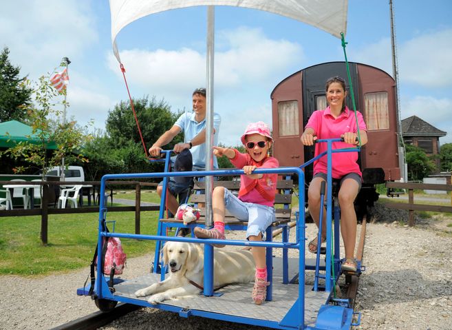 Eine Familie mit einem Hund fährt auf einer Draisine auf Schienen. Im Hintergrund sind ein alter Zugwaggon und ein Garten mit Tischen zu sehen.
