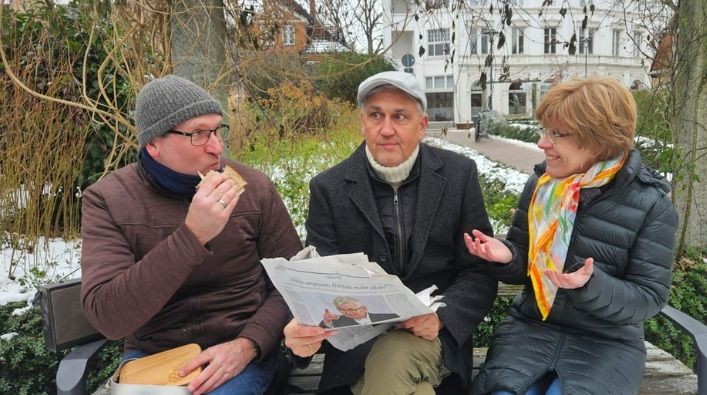 Drei Personen sitzen auf einer Parkbank im Winter. Eine Person isst ein Sandwich, eine andere hält eine Zeitung, und die dritte spricht gestikulierend.