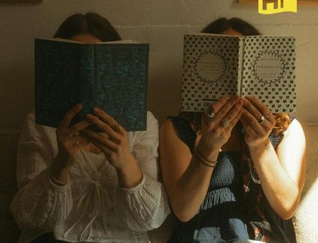 Zwei Personen sitzen auf einem Sofa und lesen Bücher, die ihre Gesichter verdecken. Ein gelbes Logo mit den Buchstaben 'HF' ist oben rechts zu sehen.