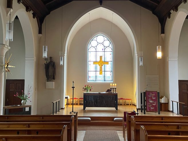 Innenansicht einer Kirche mit Altar, Bänken und einem großen Kreuzfenster im Hintergrund.