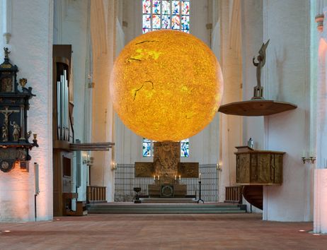 Innenansicht einer Kirche mit einer großen, leuchtenden Kugel, die die Sonne darstellt, über dem Altar.