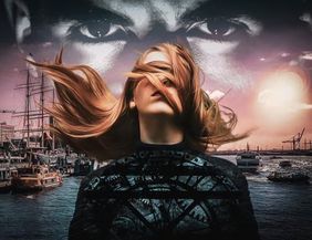 Eine Frau mit wehenden Haaren steht vor einem Hafen bei Sonnenuntergang, über ihr ein großes, geheimnisvolles Augenpaar am Himmel. Die Stimmung ist düster und emotional.