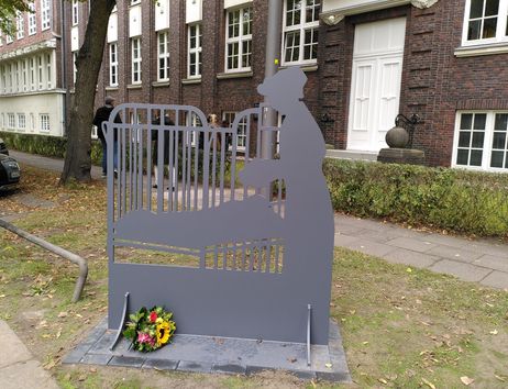 Metallskulptur eines Kinderbetts mit Krankenschwester vor einem Backsteingebäude.