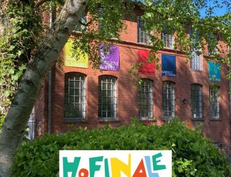 Backstein-Gebäude mit bunten Bannern und einem Schild mit der Aufschrift 'HoFinale - Wir machen Kunst!' im Vordergrund.