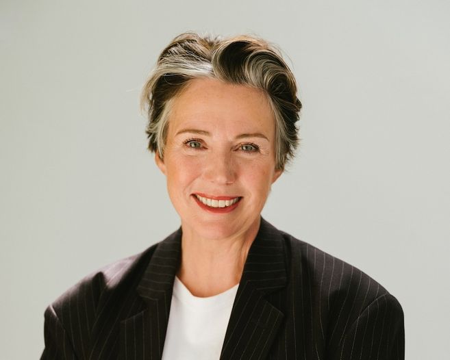 Porträt einer lächelnden Frau mit kurzem Haar in einem dunklen Anzug vor hellem Hintergrund.