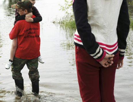 Ein Junge trägt ein kleineres Kind durch flaches Hochwasser, während eine Person im Vordergrund zuschaut.