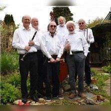 Eine Jazzband mit sechs älteren Männern in weißen Hemden posiert im Freien mit ihren Instrumenten.