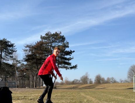 Ein Mann in roter Sportkleidung spielt Discgolf auf einem offenen Feld mit Bäumen im Hintergrund.