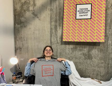 Foto einer Nähwerkstatt mit Nähmaschine im Vordergrund und junger Frau, die lächelnd ein graues T-Shirt hochhält auf dem "Fashion Revolution Germany" steht.  